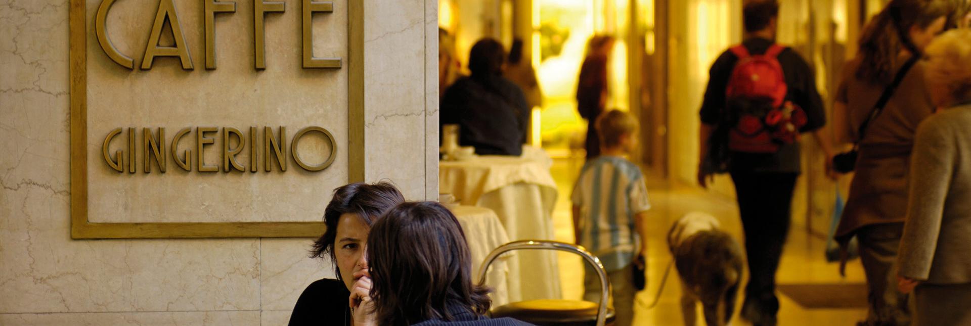 People chatting outside Caffè Gingerino in Bozano, with shoppers walking through the Lauben in the background.