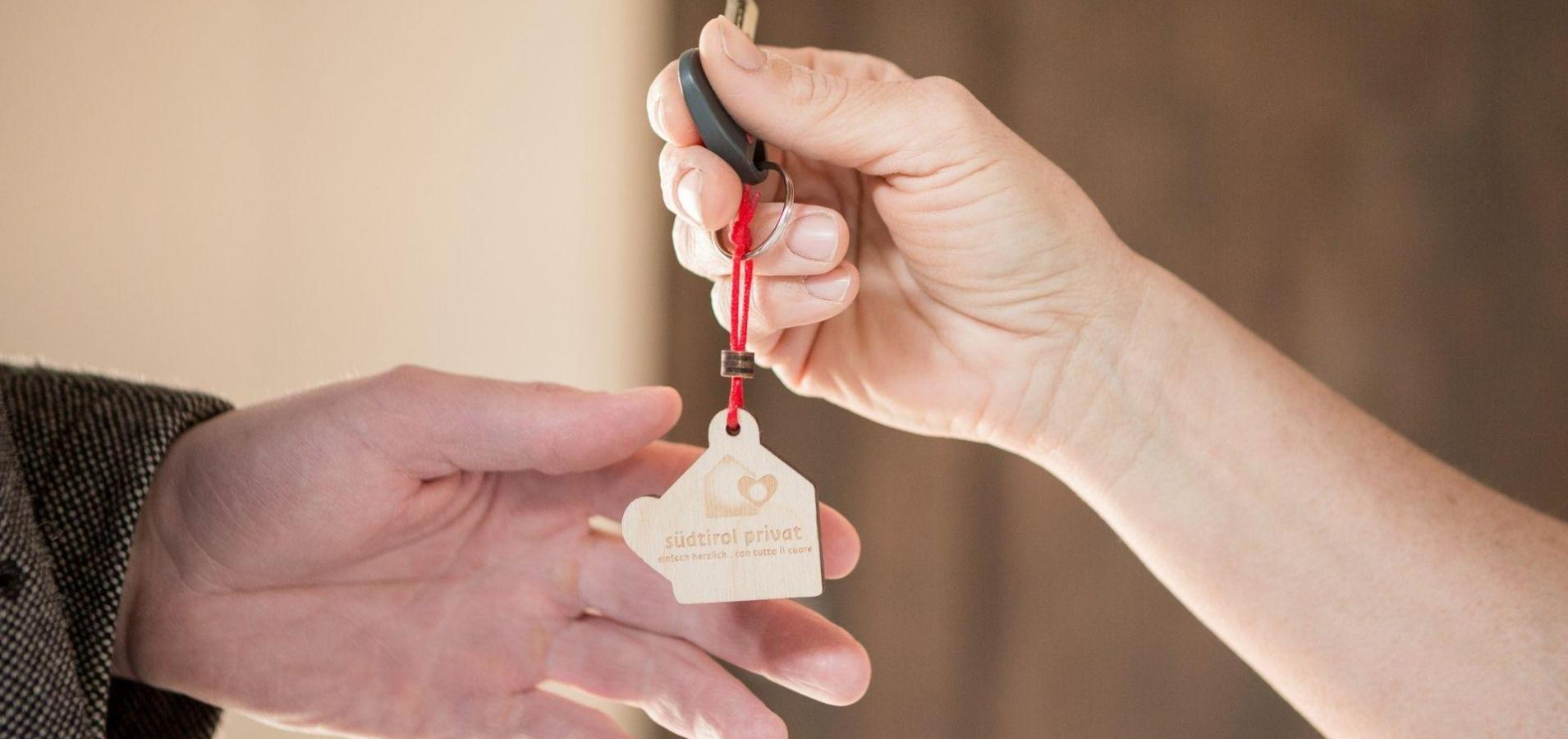 Schlüsselübergabe mit Holzanhänger „südtirol privat“ zwischen zwei Händen in Nahaufnahme.