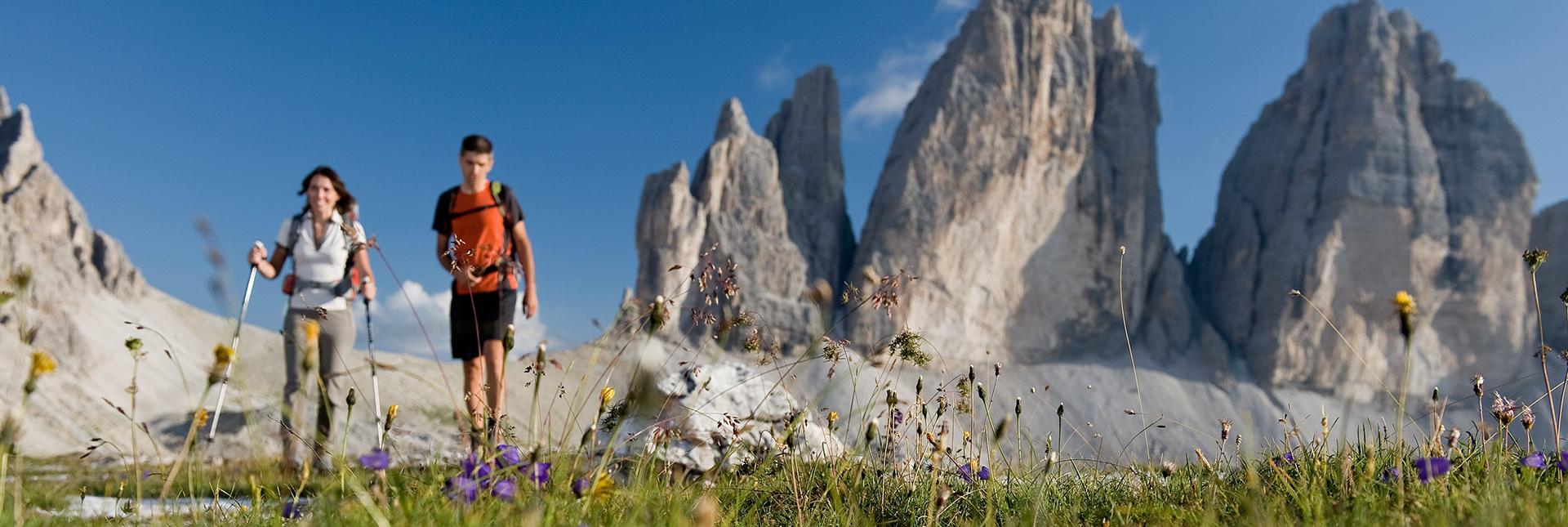 Due escursionisti camminano su un prato fiorito davanti alle Tre Cime sotto un cielo azzurro.
